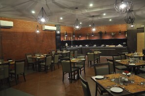 Restaurante com várias mesas e cadeiras, área de cozinha e luminárias pendentes.