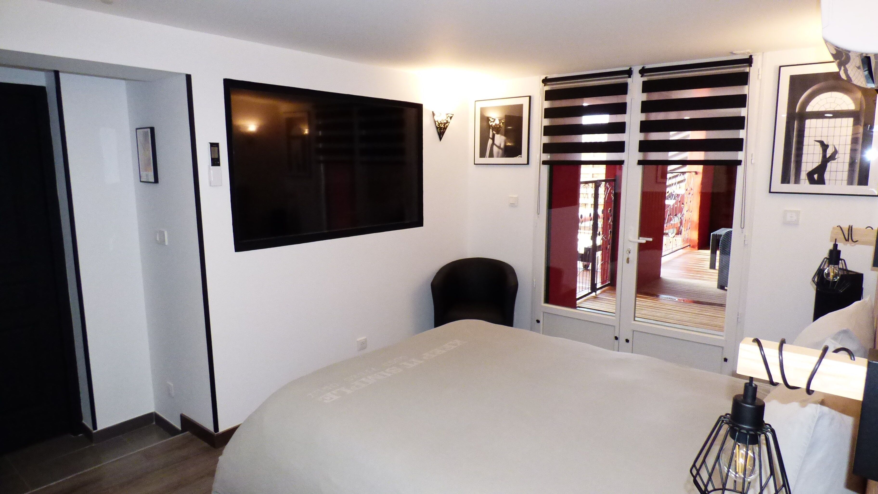 Habitación de hotel con cama, una silla, un televisor montado en la pared, una puerta y una ventana con persianas.