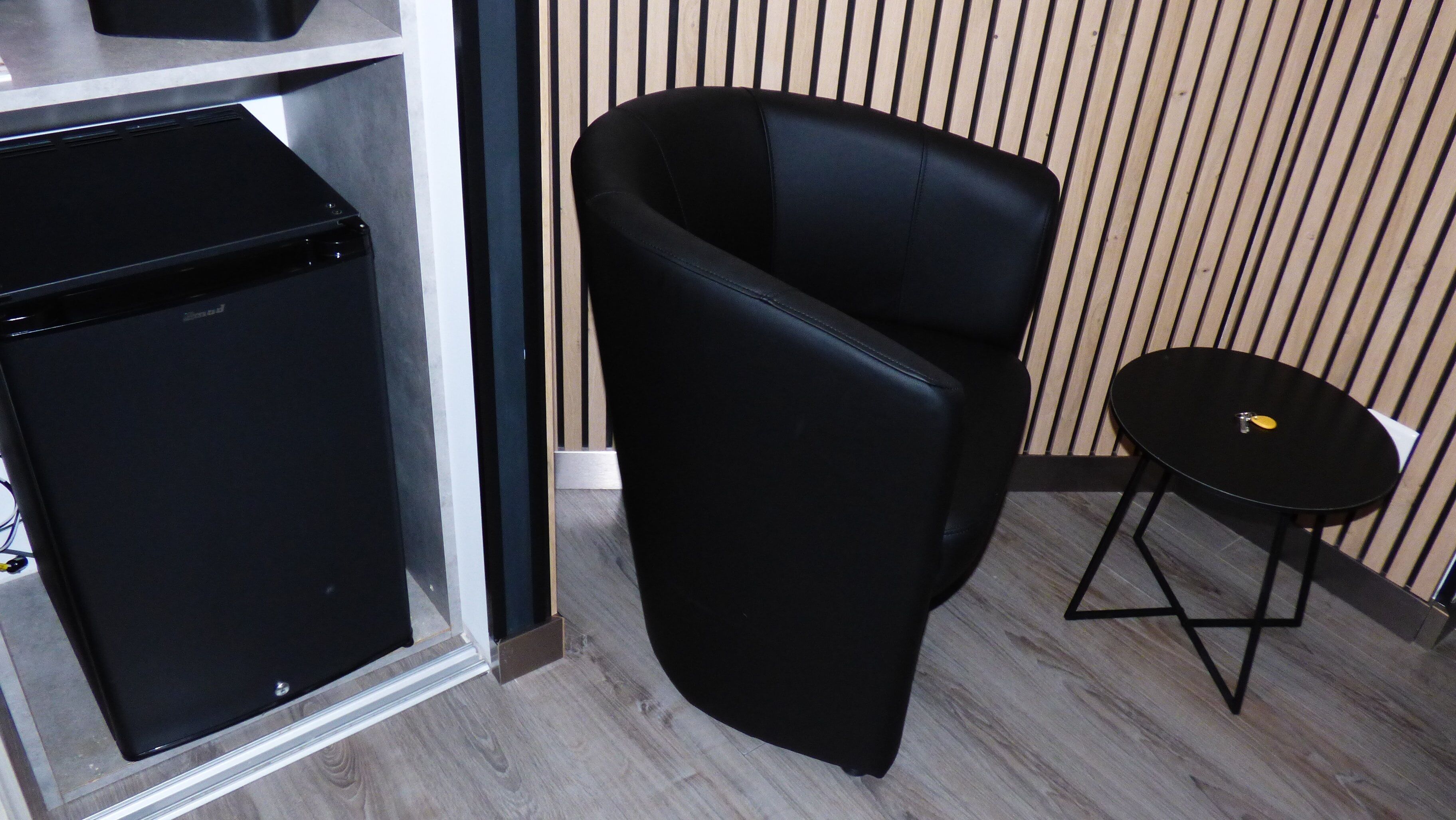 A compact room with a black refrigerator, a black chair, and a small round table.