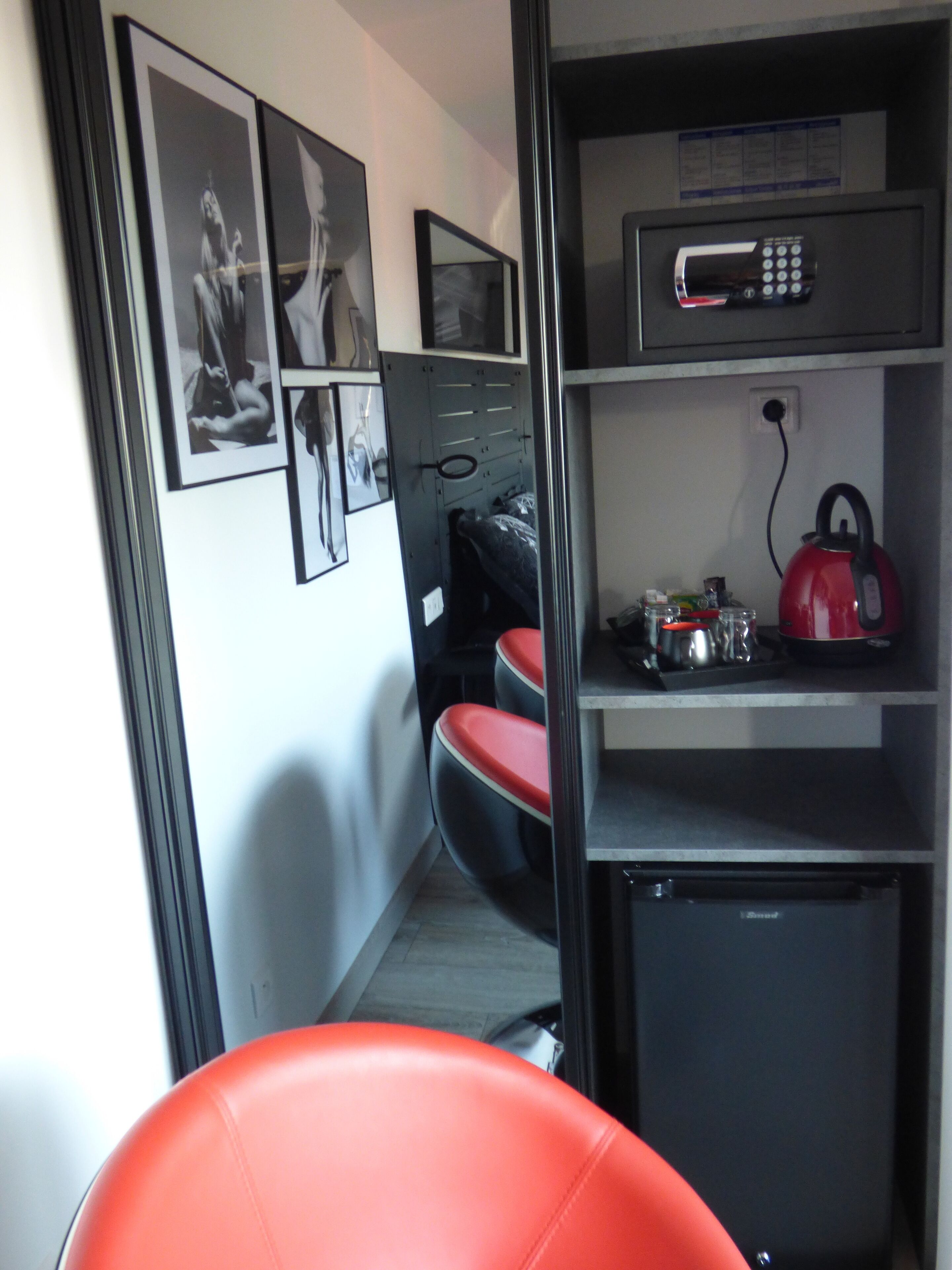 A compact room with a kitchenette, a microwave, a kettle, and a small refrigerator. There are framed pictures on the wall and a red chair in the foreground.
