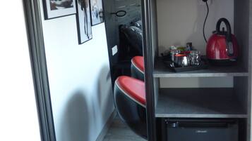 A compact room with a kitchenette, a microwave, a kettle, and a small refrigerator. There are framed pictures on the wall and a red chair in the foreground.