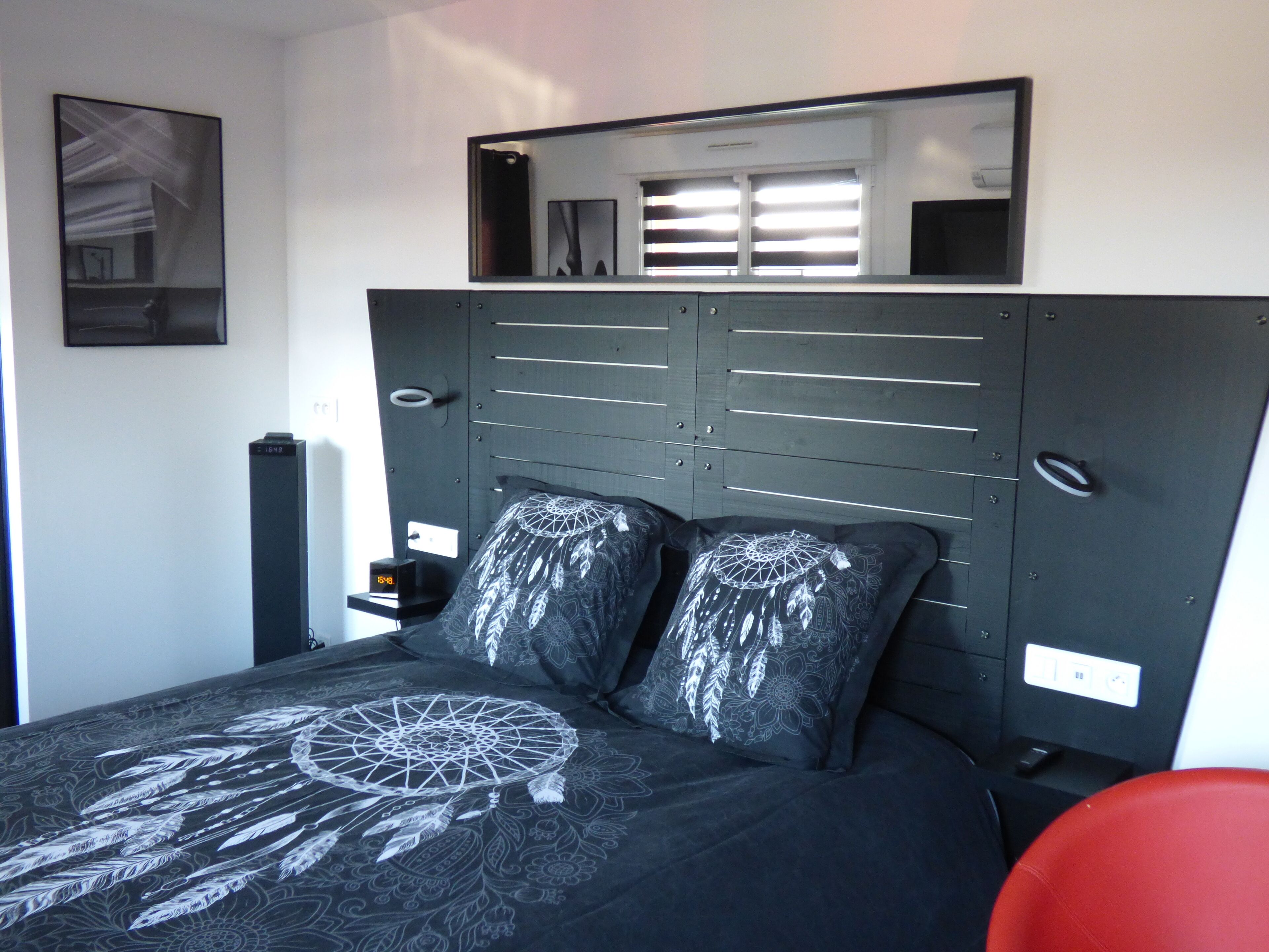 Une chambre à coucher avec un lit, une table de chevet, un fauteuil rouge, un miroir fixé au mur et un tableau encadré.