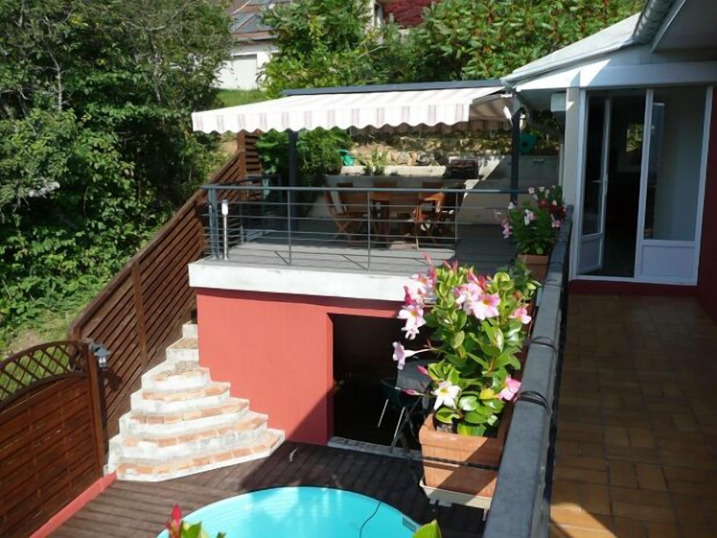 Un espace piscine avec une terrasse en bois, un escalier menant à un espace de détente extérieur abrité, et une vue sur un immeuble d’habitation en arrière-plan.