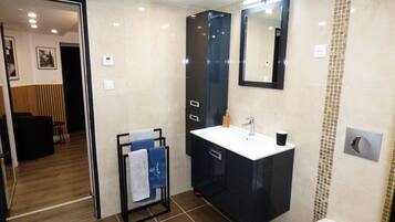 A modern bathroom with a black vanity, white sink, and a large mirror. There is a towel rack with towels, a black chair, and a wooden bench in the background.