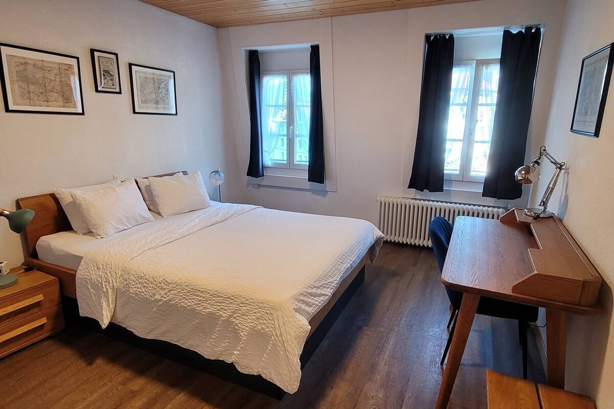 Um quarto com cama, escrivaninha, cadeira, mesa de cabeceira e radiador.