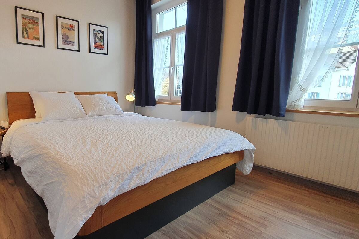 Cama bem arrumada com edredom branco, cabeceira de madeira e mesa de cabeceira com abajur. Há três quadros na parede acima da cama.