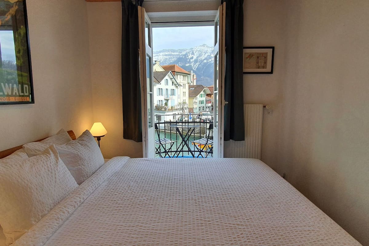 Quarto de hotel com cama, luminária, quadro e vista para edifícios e montanhas por uma porta aberta.