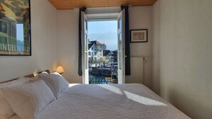 Habitación de hotel con cama, ventana con vista a edificios y montañas, un cuadro en la pared y una lámpara en la mesita de noche.