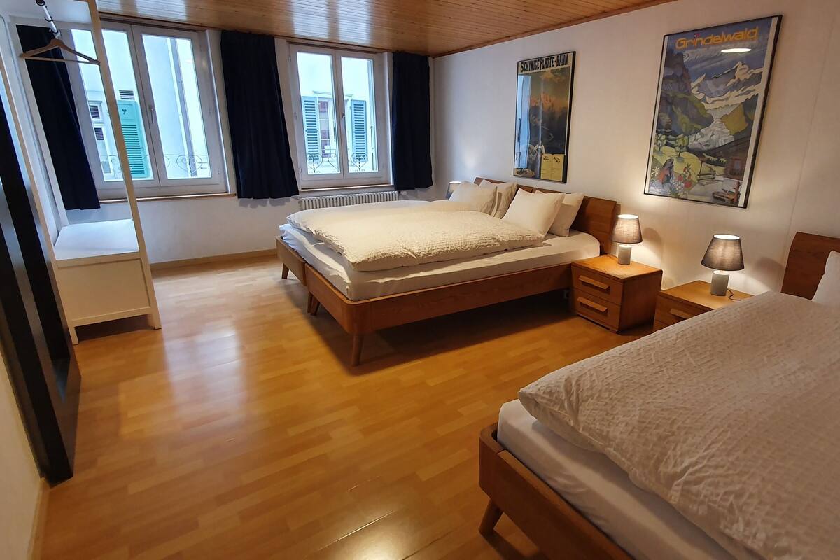 Quarto com duas camas, piso de madeira e janelas amplas com cortinas.