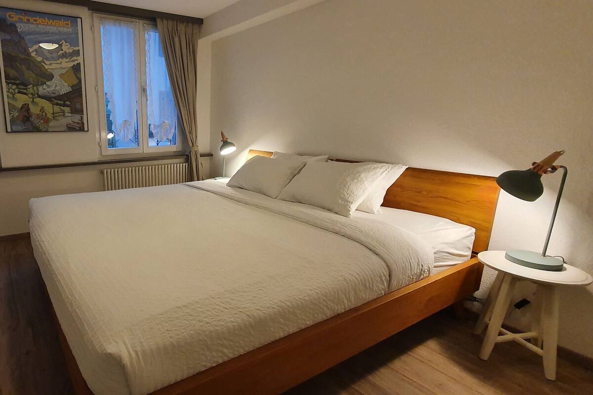 Uma cama bem arrumada com cabeceira de madeira, uma mesinha de cabeceira com abajur e um cartaz na parede.