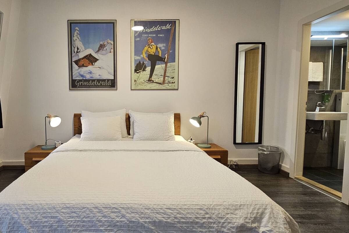 Quarto de hotel com uma cama grande, duas mesas de cabeceira com luminárias, uma janela com cortinas, um pôster na parede e um espelho no banheiro.