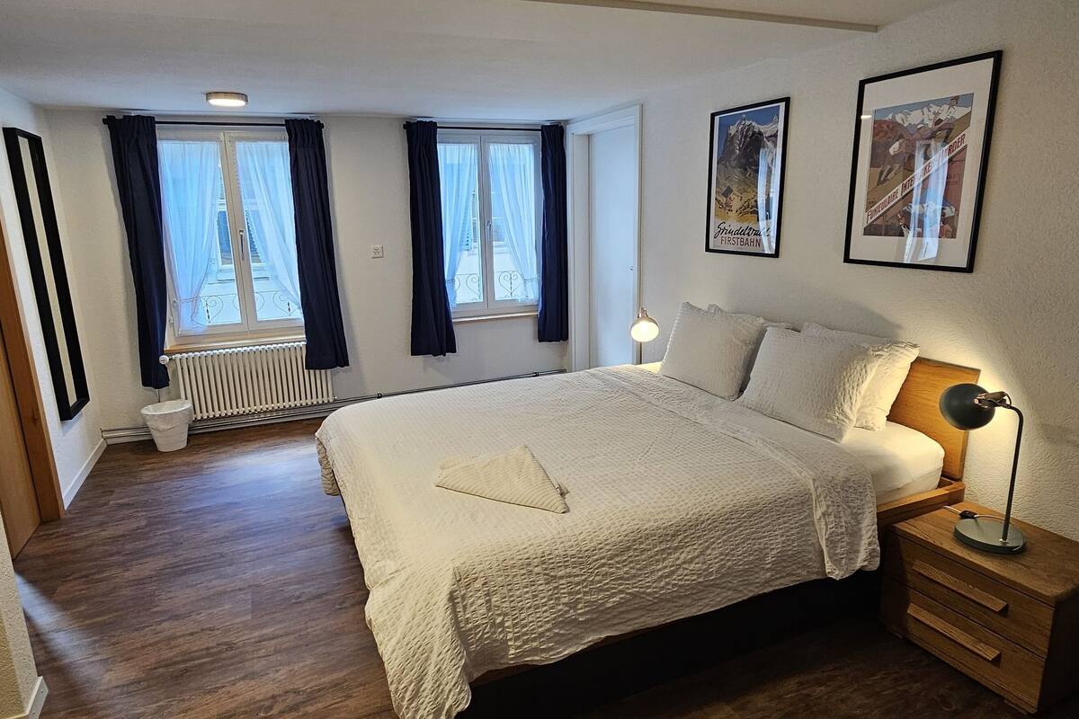 Quarto com uma cama grande, duas janelas com cortinas, um radiador, abajur e quadros nas paredes.