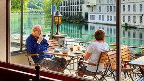 Un café con amplios ventanales que dan a un canal, con mobiliario de madera y vistas de edificios al otro lado del agua.