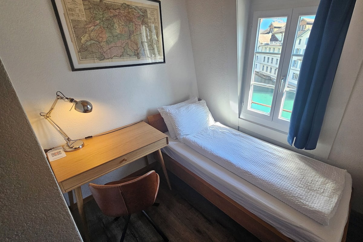 Quarto pequeno com uma cama de solteiro, uma escrivaninha e uma cadeira, um mapa na parede e uma janela com vista para edifícios.