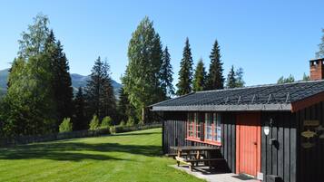 A rustic wooden cabin with a red door, surrounded by a grassy area and tall trees.
