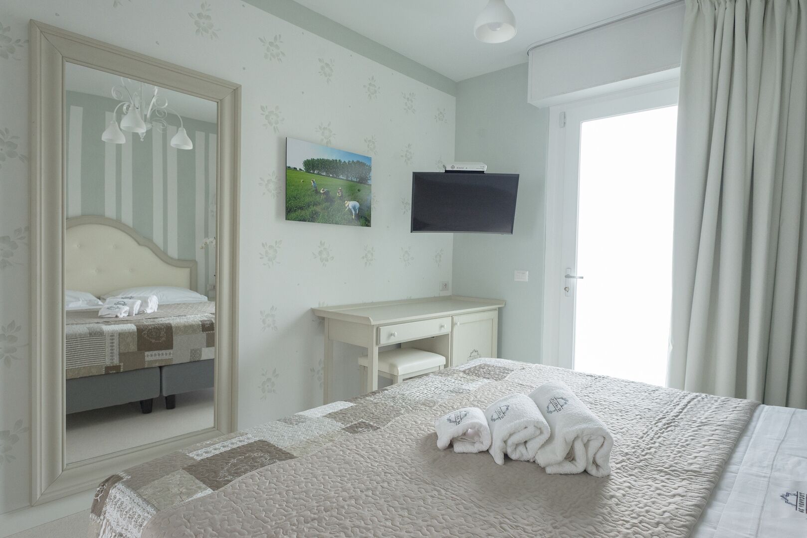 A bedroom with a bed, a desk, a mirror, a TV, and a door.