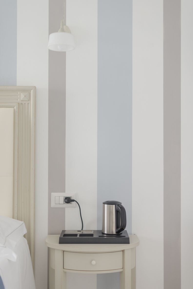 A hotel room with a bed, a bedside table with a kettle, a lamp, and a power outlet.