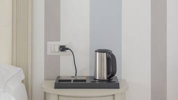 A hotel room with a bed, a bedside table with a kettle, a lamp, and a power outlet.