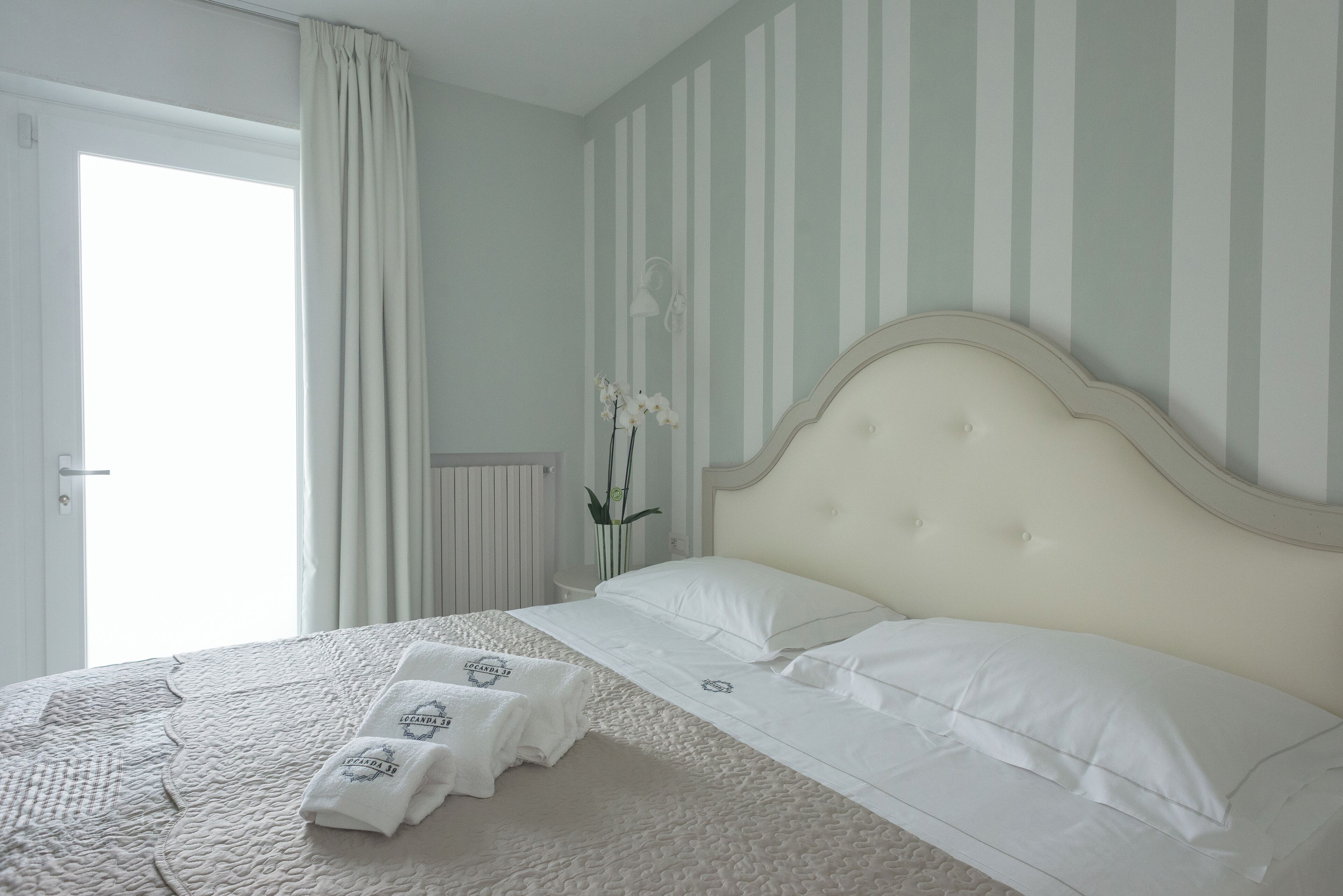 A neatly made bed with white linens and pillows, a bedside table with a vase of flowers, and a window with curtains.