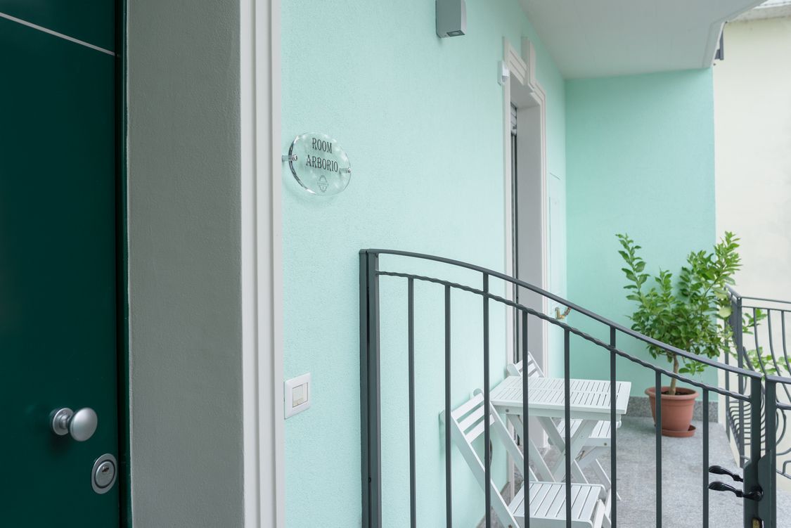 Una stanza con un cartello che indica 'ROOM ARBORIO', un tavolo bianco, una pianta in vaso e una ringhiera metallica.