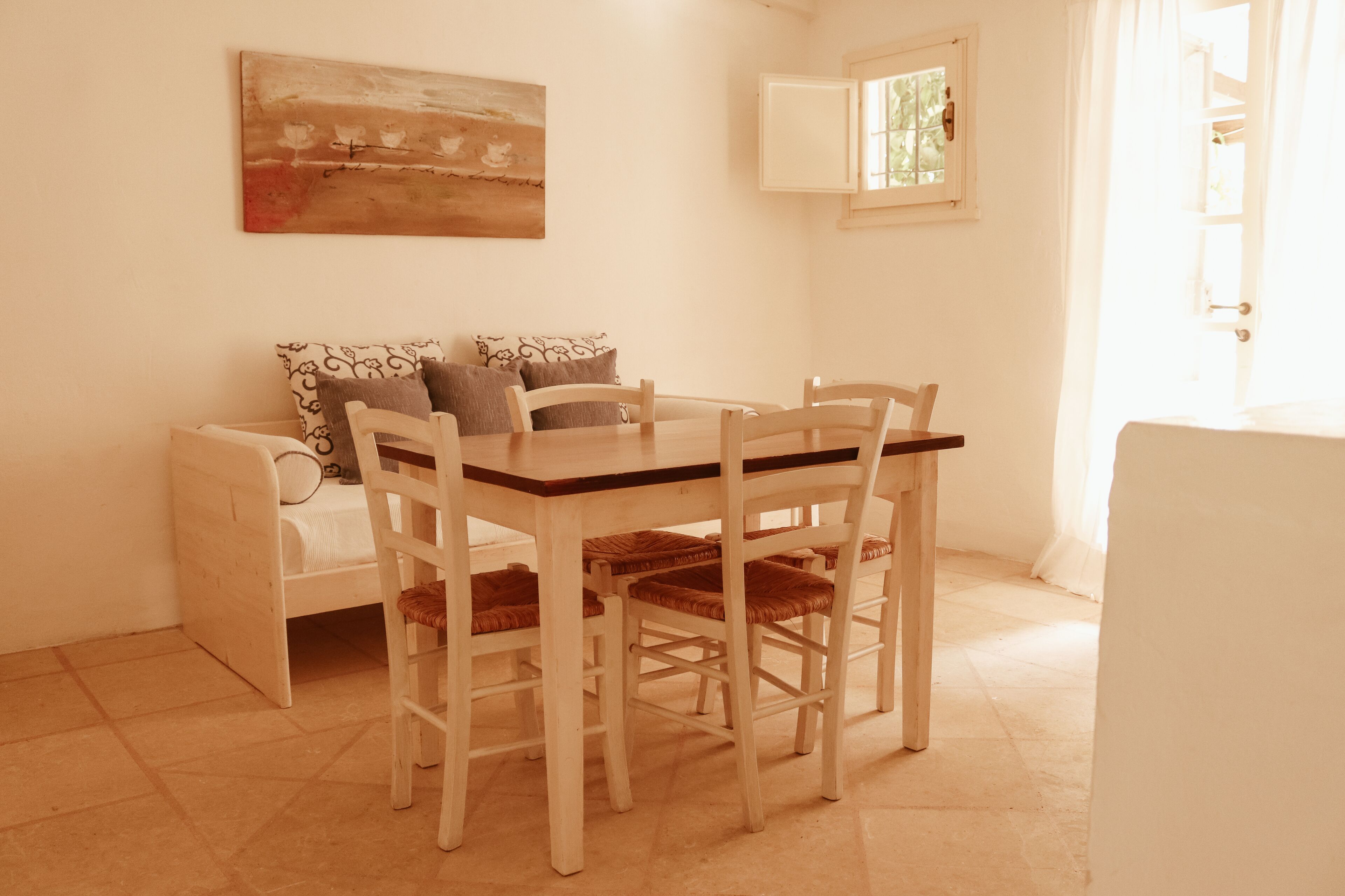A dining area with a wooden table and chairs, a sofa with cushions, a painting on the wall, and a window with curtains.