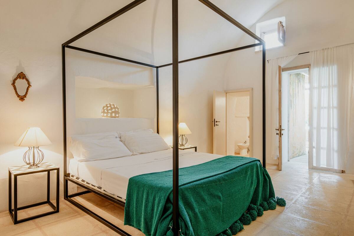 A four-poster bed with a green bedspread, two bedside lamps, and a view of the outdoors through a door.