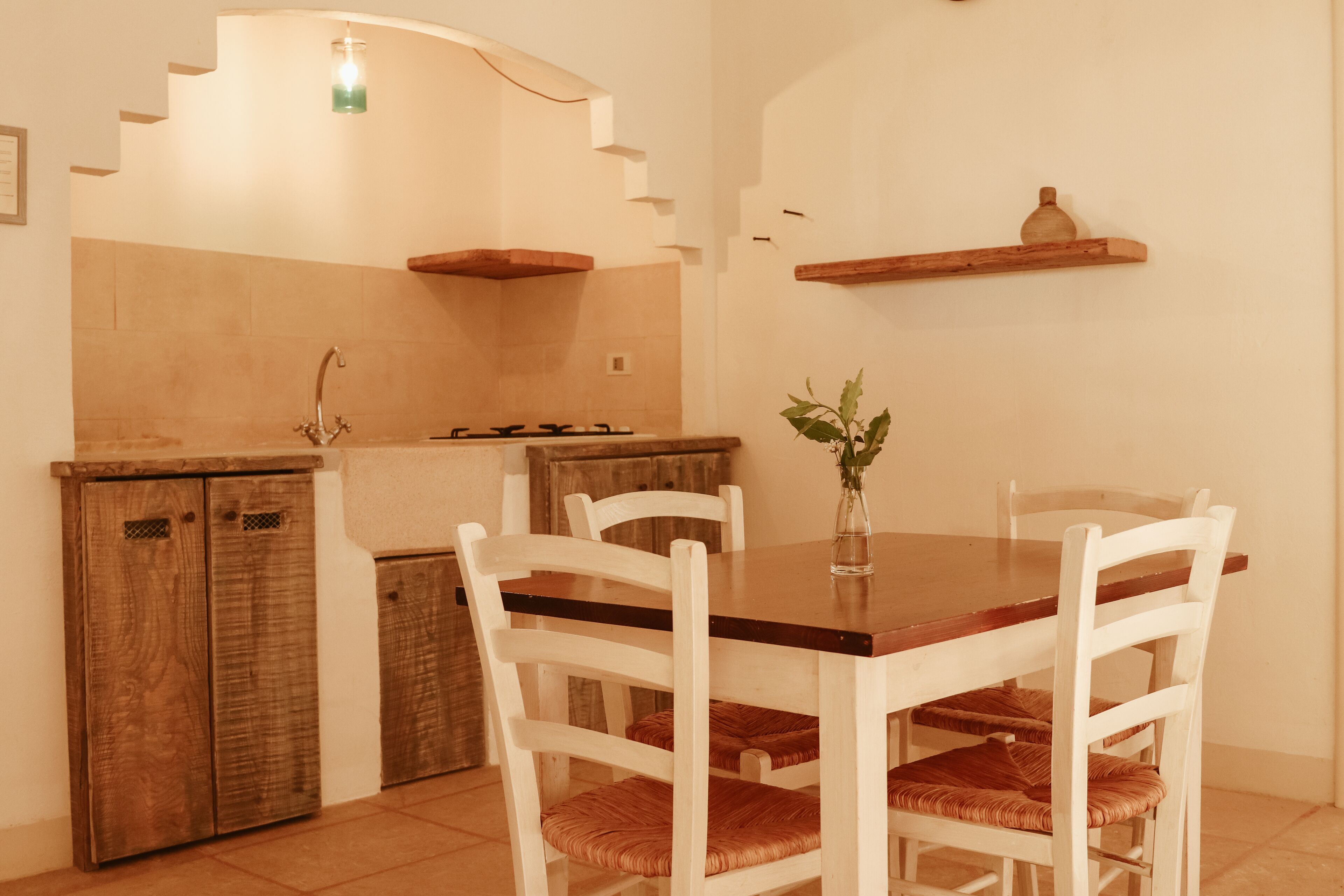 Une cuisine avec une table en bois, des chaises blanches, un luminaire suspendu et une petite étagère.