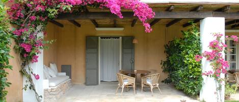 A covered patio with a dining table and chairs, a sofa, and a stone pathway leading to a door.