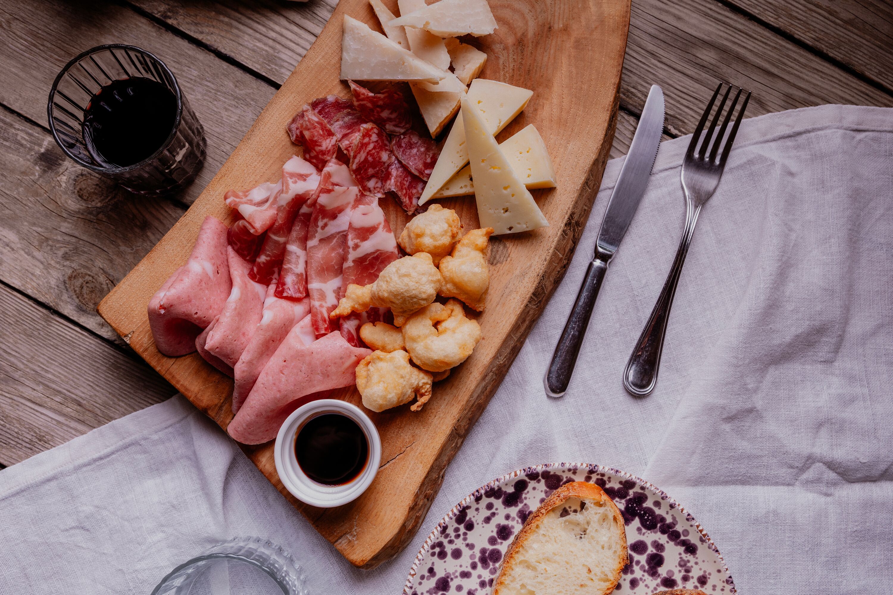 A wooden board with various cheeses, cured meats, and fried items, accompanied by a small bowl of sauce, on a rustic wooden table.