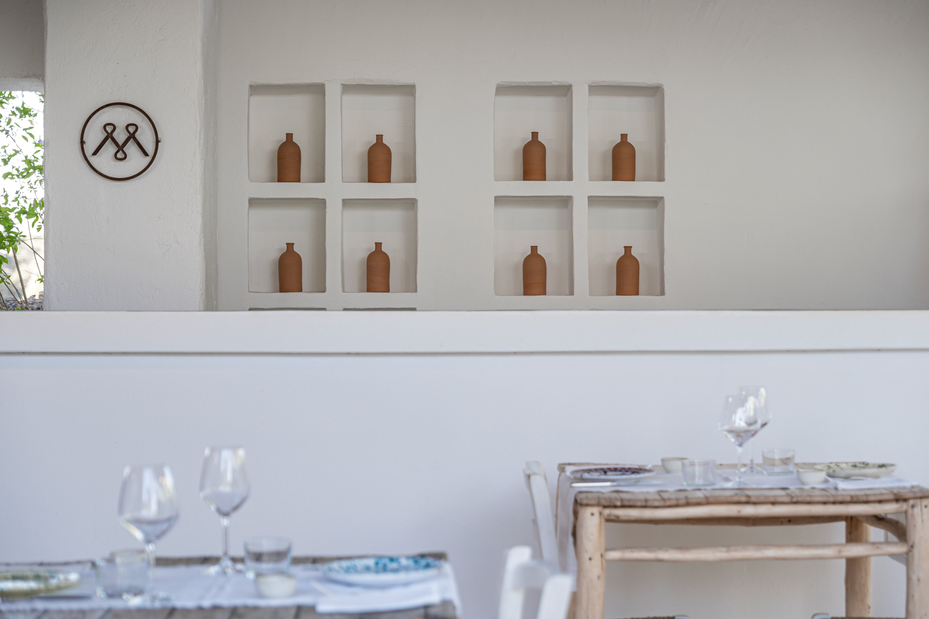 A dining area with a table set for two, a white wall with shelves containing ceramic vases, and a circular logo on the wall.