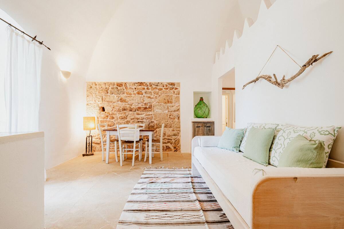 A cozy living room with a stone wall, a wooden sofa, a dining table with chairs, and a hanging branch decoration.