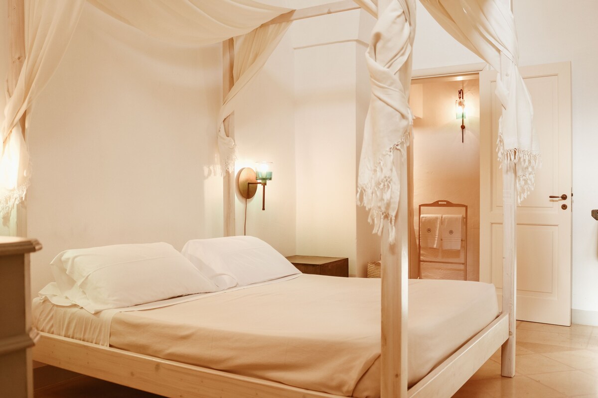 A four-poster bed with a canopy, a bedside table, a wall-mounted lamp, and a door leading to a bathroom.