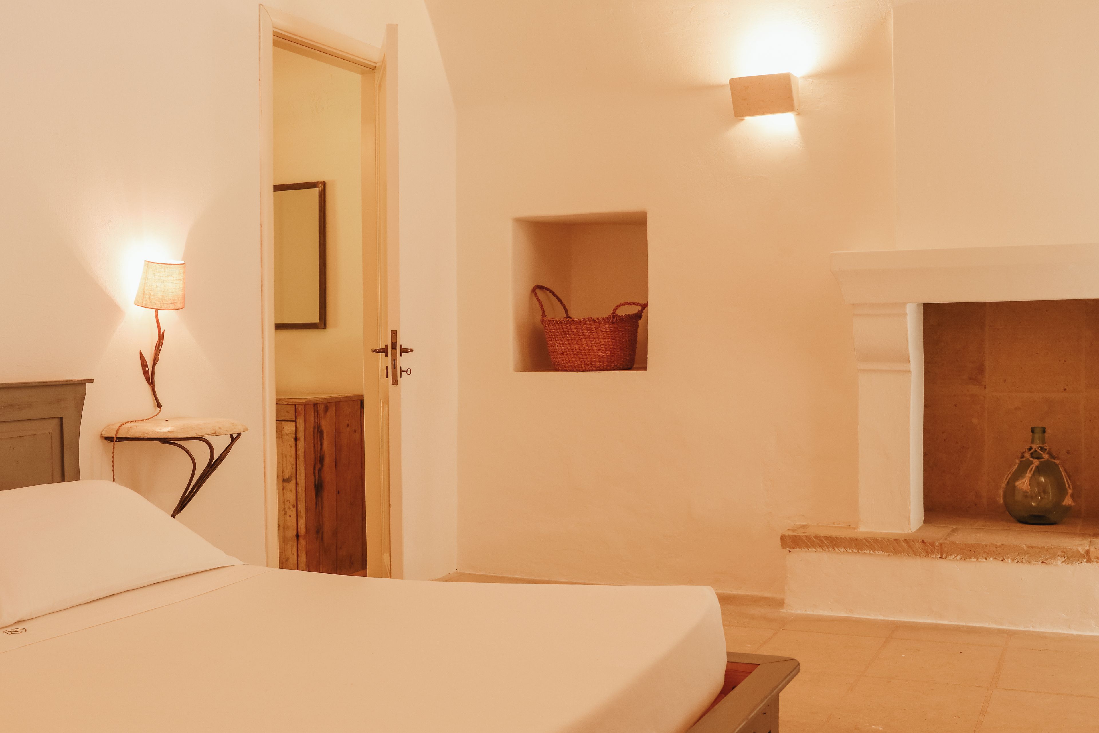 Un dormitorio con cama, mesita de noche, lámpara, espejo, cesta en la pared y chimenea.