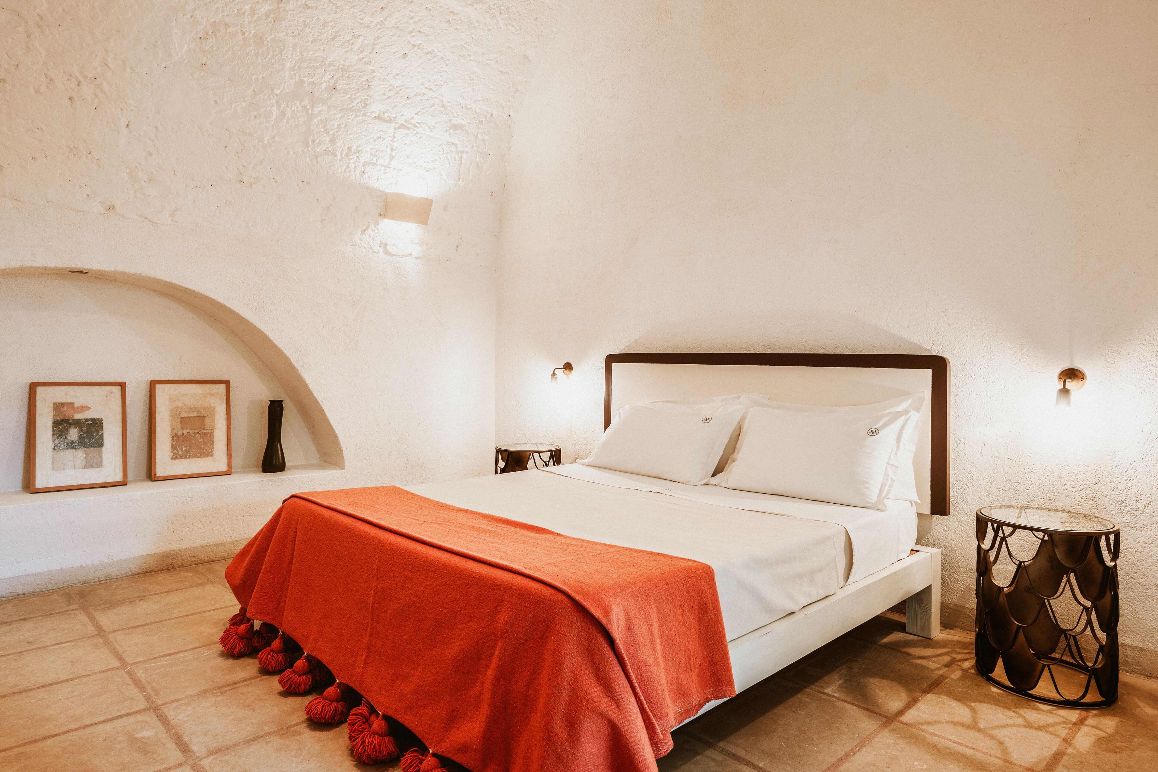 A bedroom with a bed, a red blanket, a round side table, and a wall-mounted light fixture.