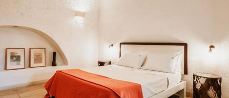 A bedroom with a bed, a red blanket, a round side table, and a wall-mounted light fixture.