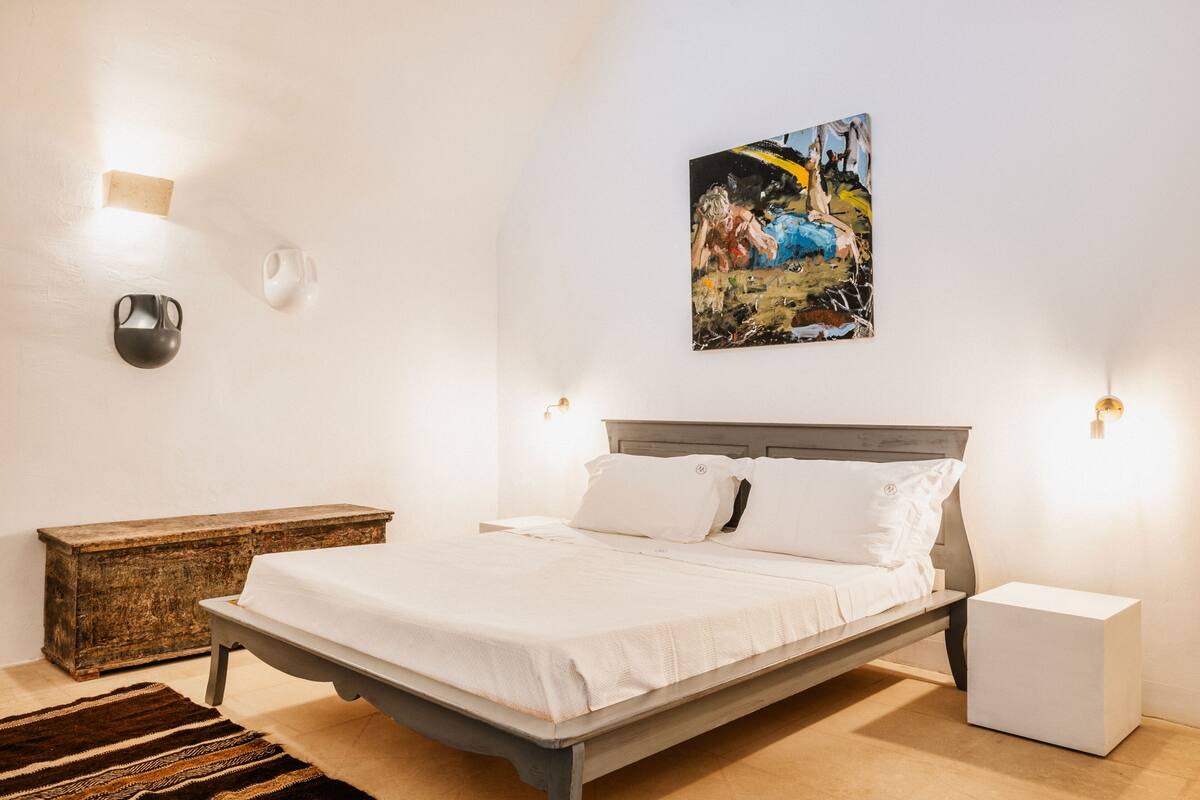 A bedroom with a bed, bedside tables, a nightstand, a painting, and a wall-mounted light fixture.