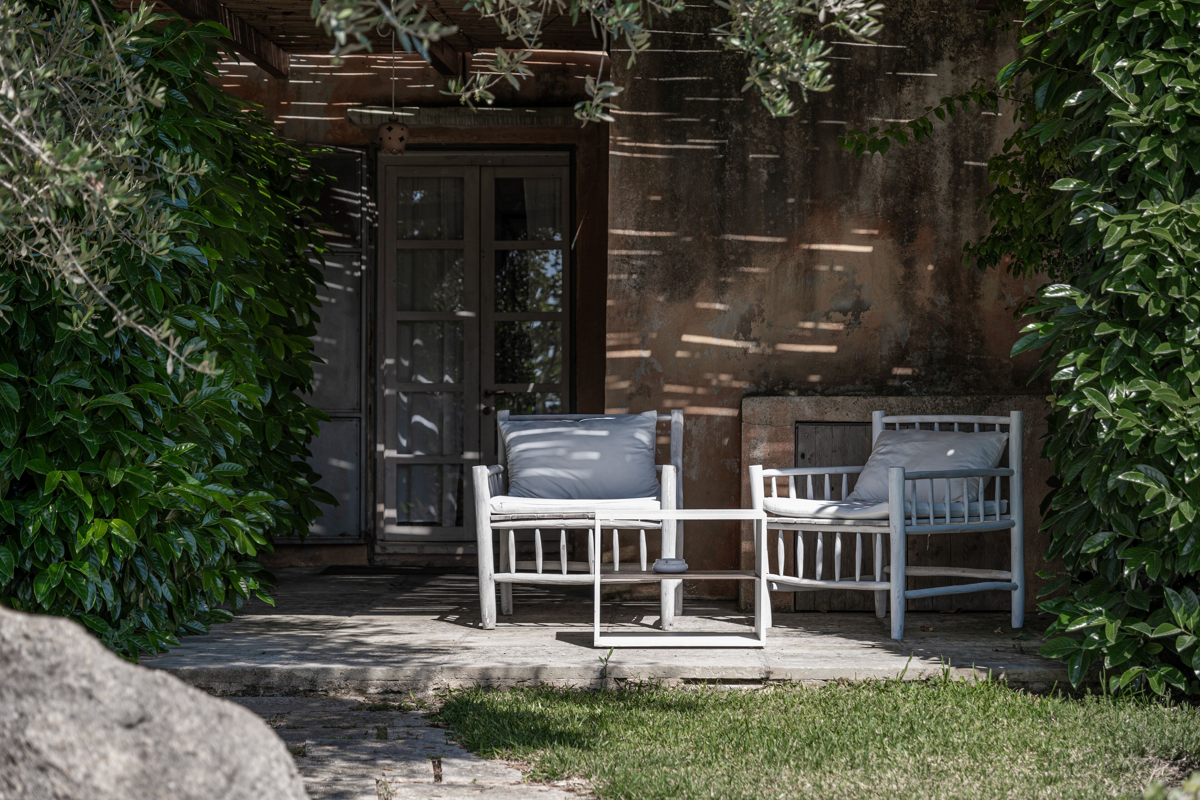 A white bench with a cushion in front of a rustic building with a wooden door.