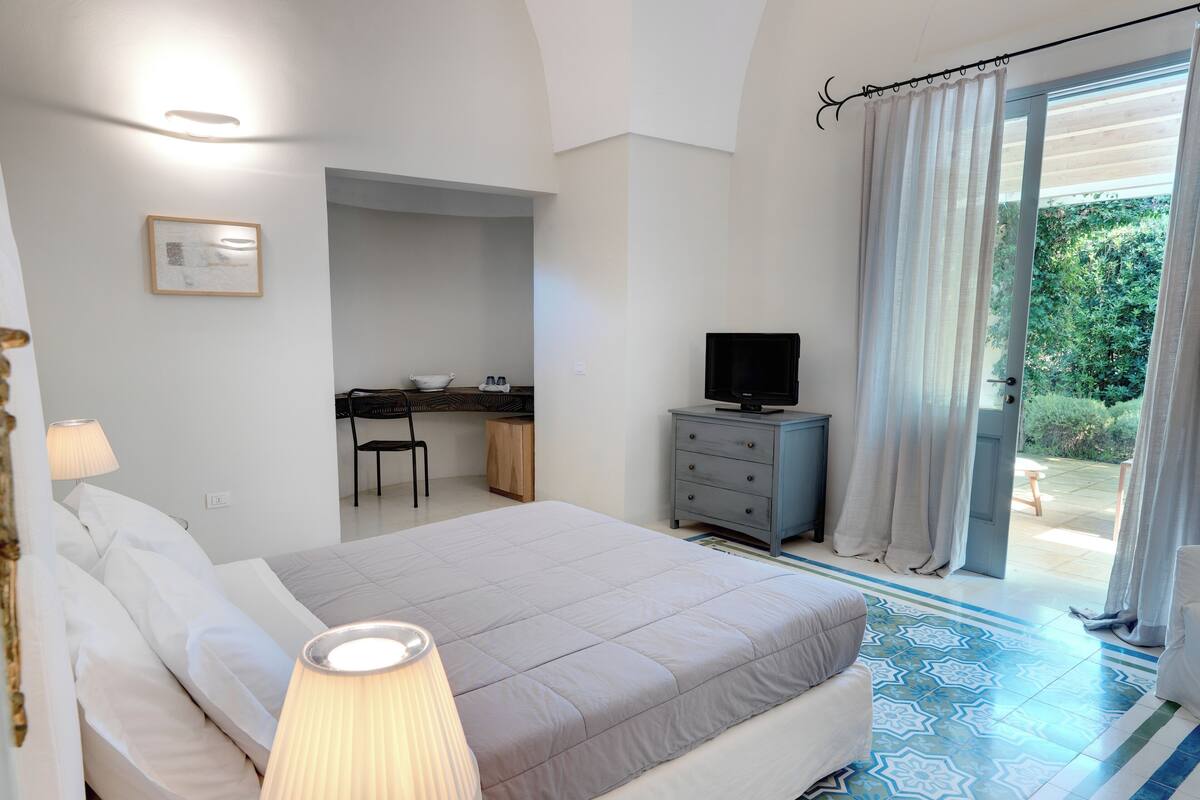 A bedroom with a bed, bedside lamp, desk, chair, and a television.