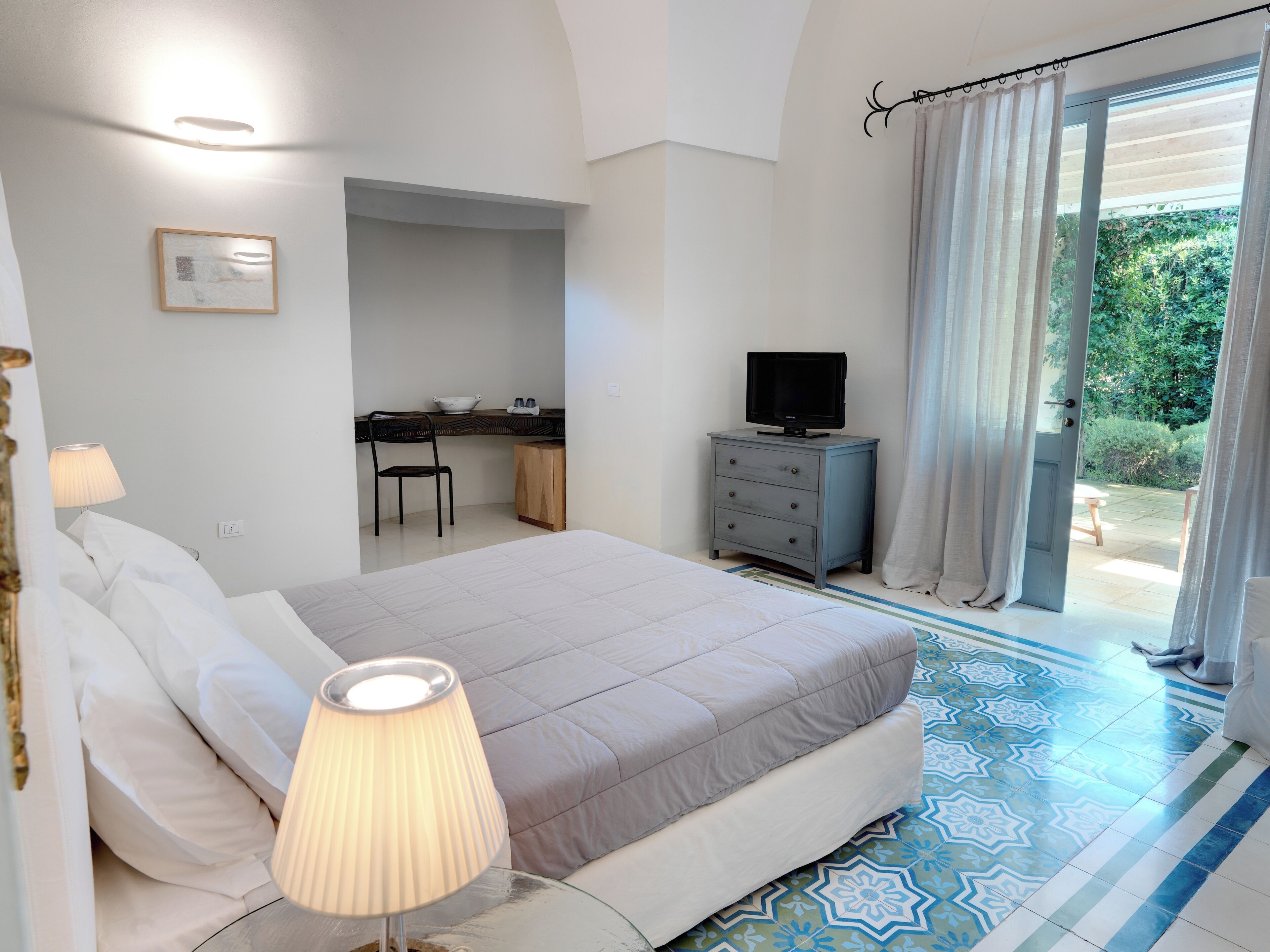 A bedroom with a bed, bedside lamp, desk, chair, and a television.