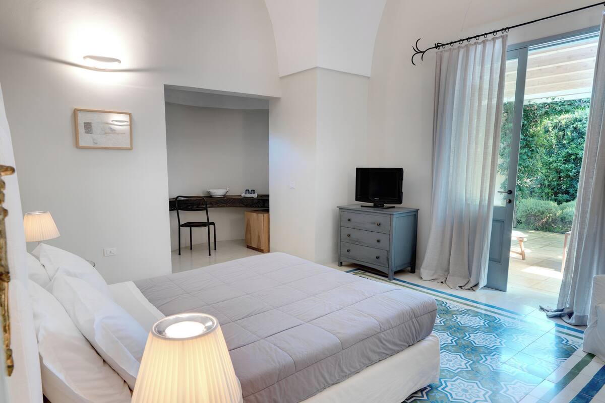 A bedroom with a bed, bedside lamp, desk, chair, and a television.