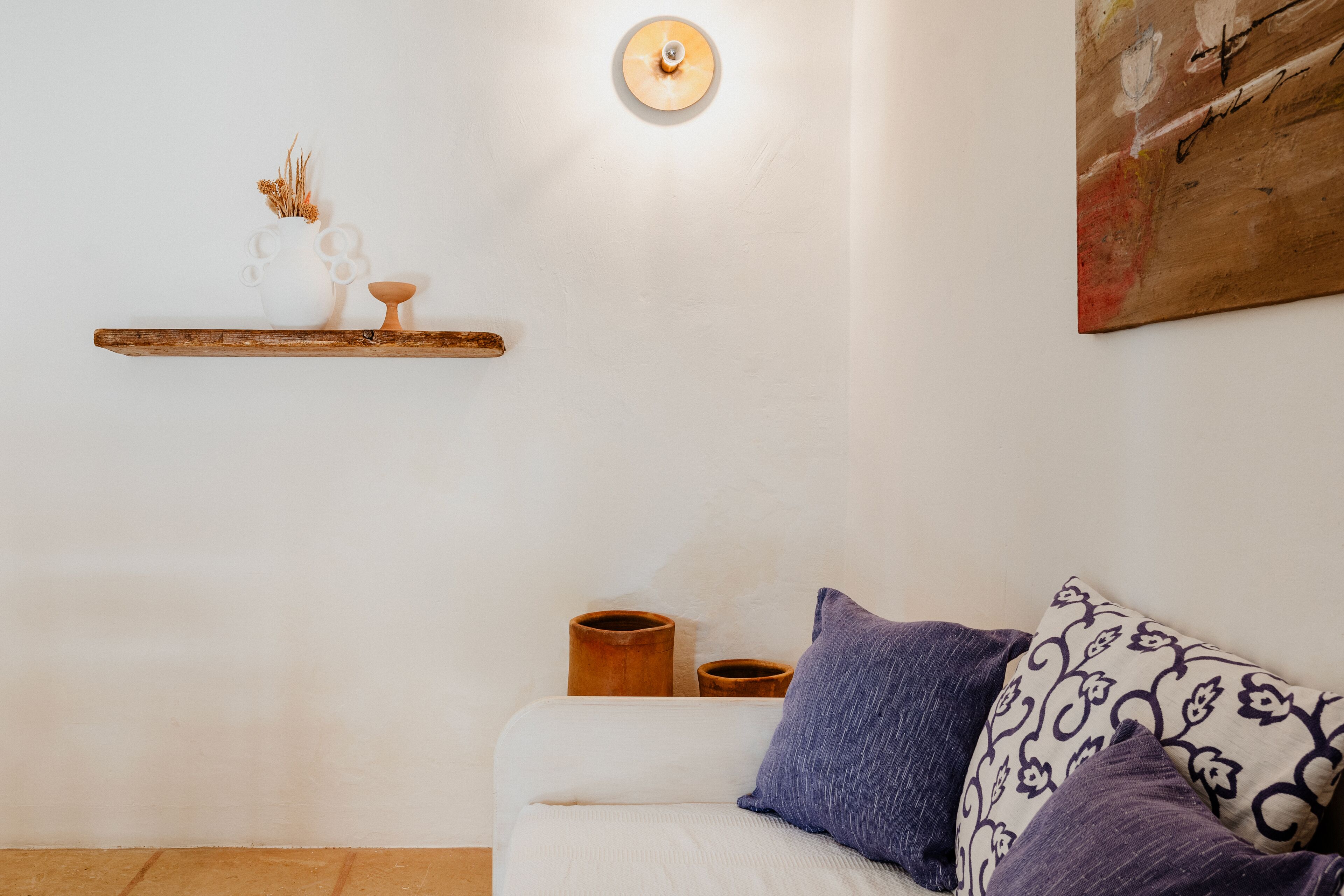 A modern living room with a white sofa, decorative pillows, a wooden shelf with small vases, and a wall-mounted light fixture.