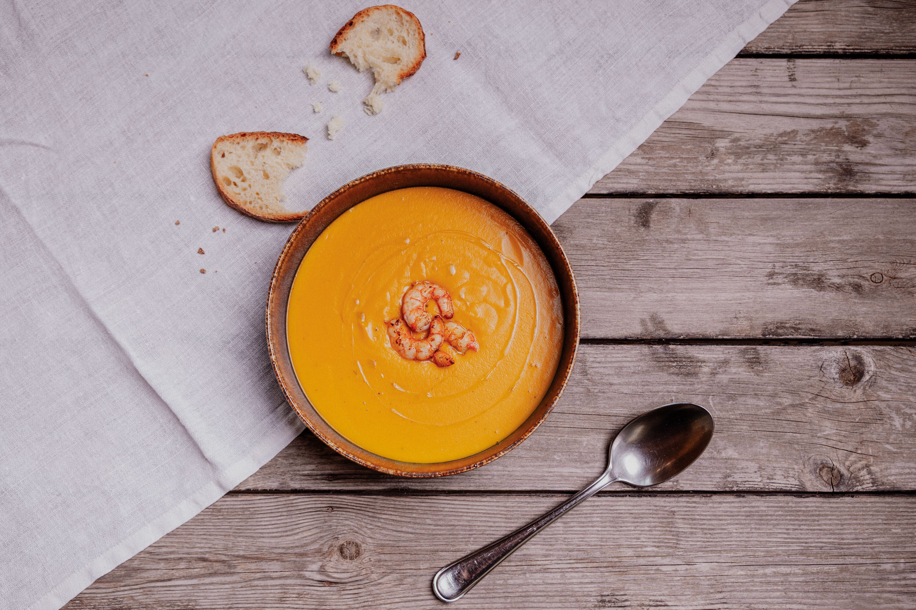 Un bol de soupe garni de crevettes, posé sur une table en bois, accompagné d’une cuillère et d’une tranche de pain.