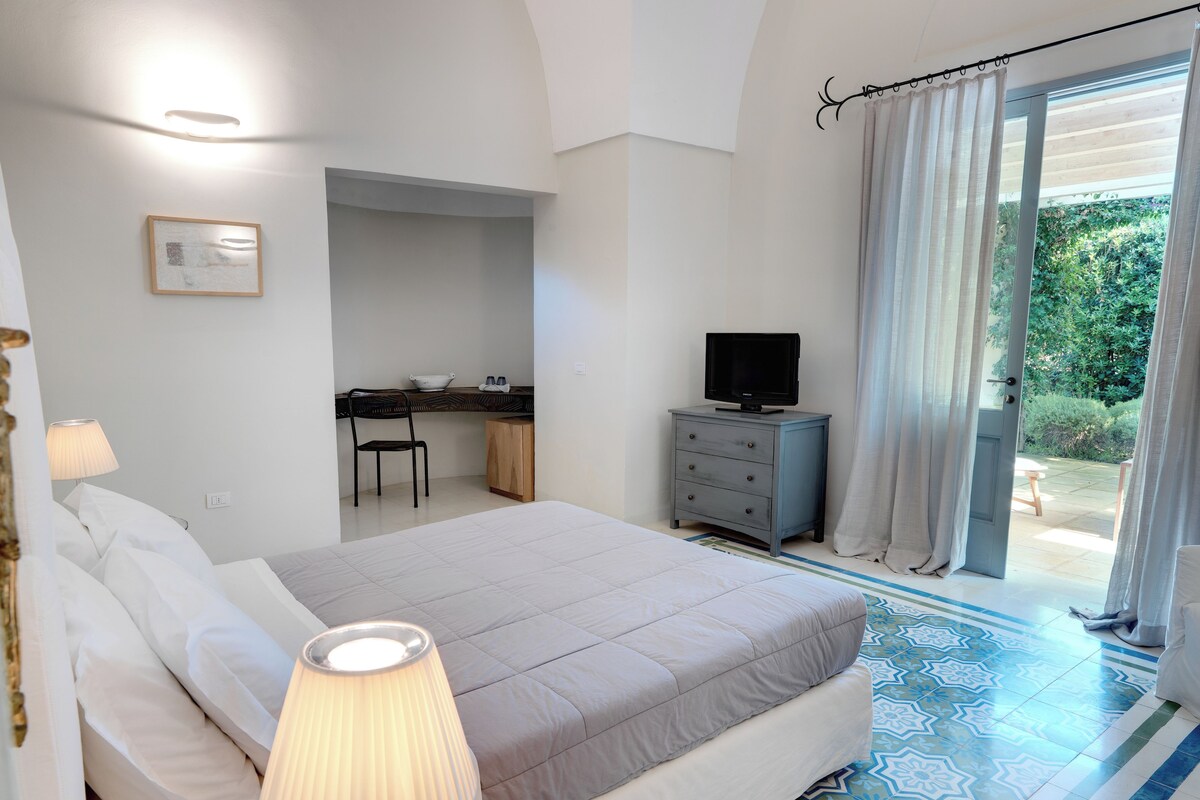 A bedroom with a bed, bedside lamp, desk, chair, and a television.