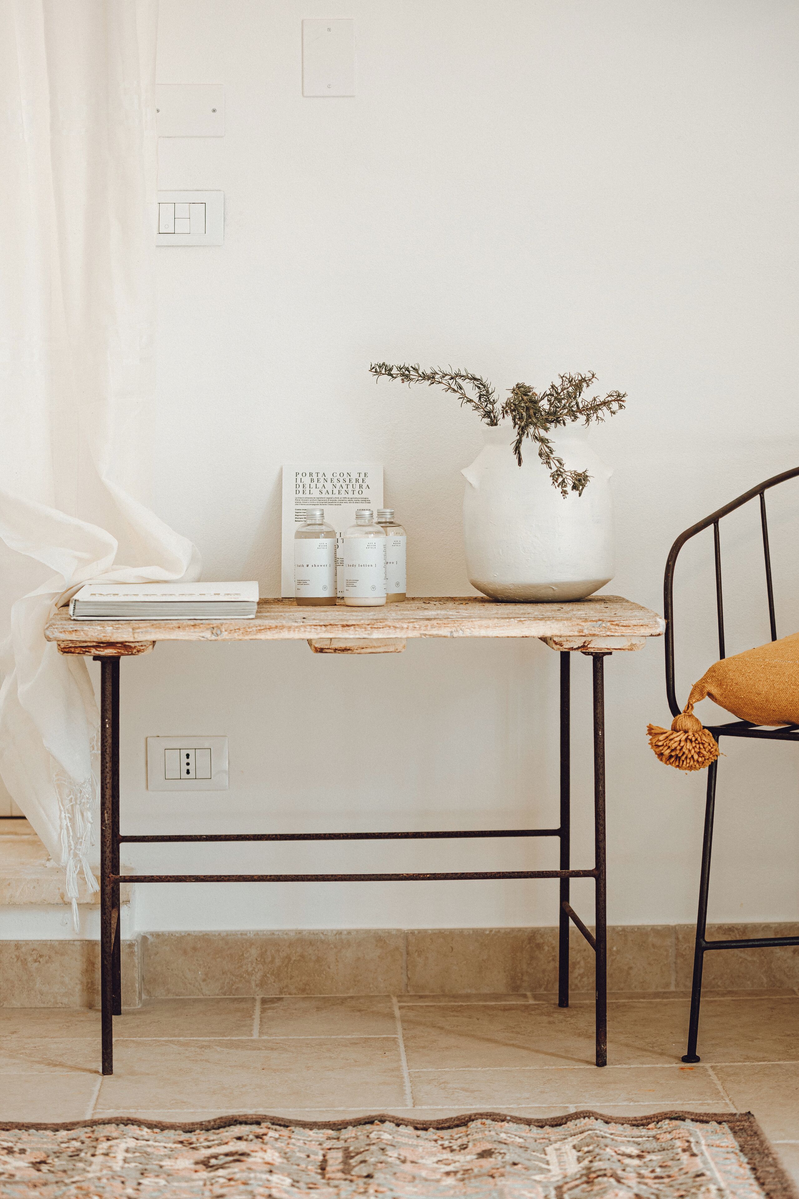 A rustic wooden table with a potted plant, a book, and two bottles, positioned next to a metal chair with a cushion.