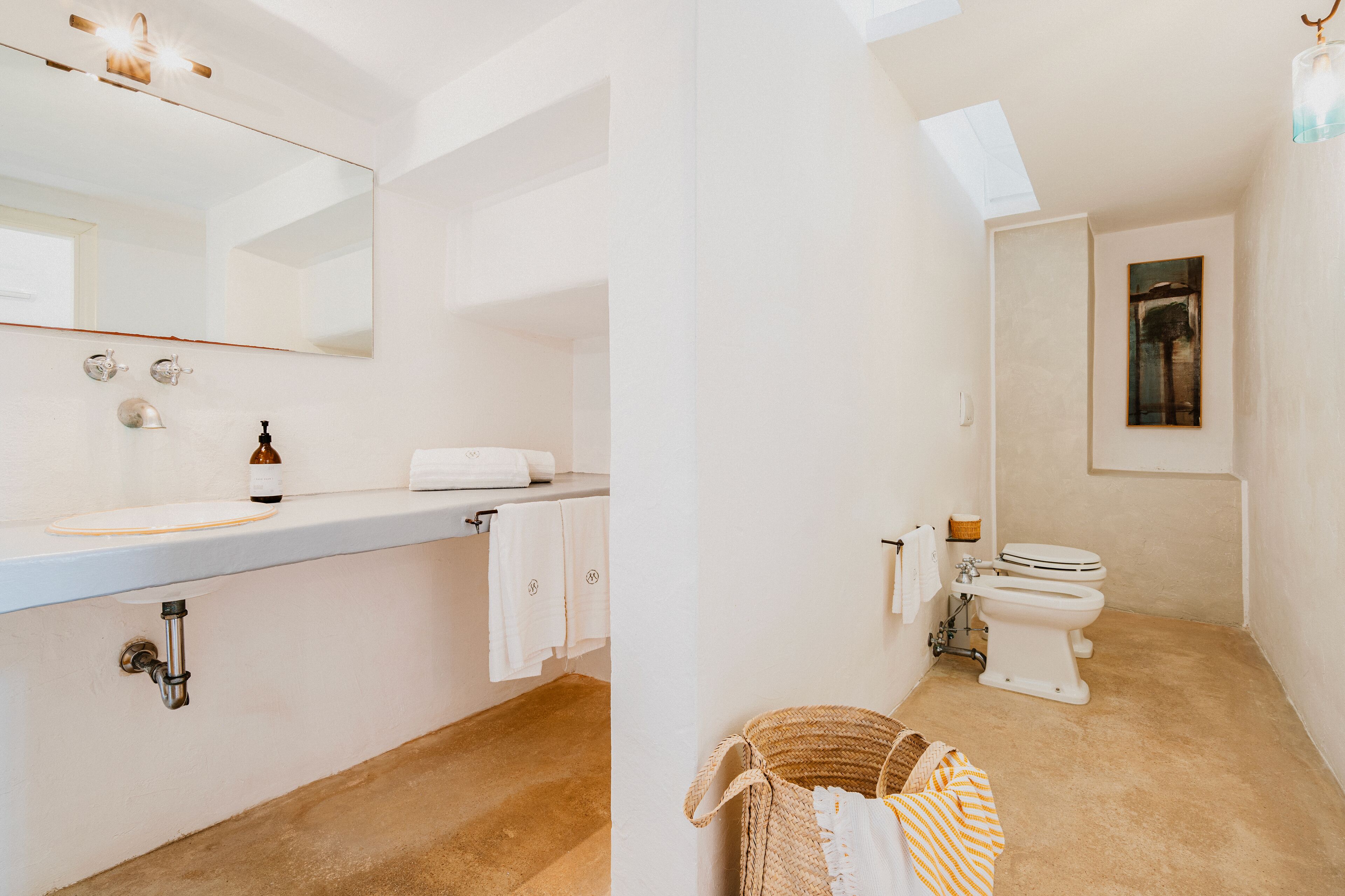 A bathroom with a white sink, mirror, and toilet. There is a towel rack with towels, a small basket, and a bottle on the sink.
