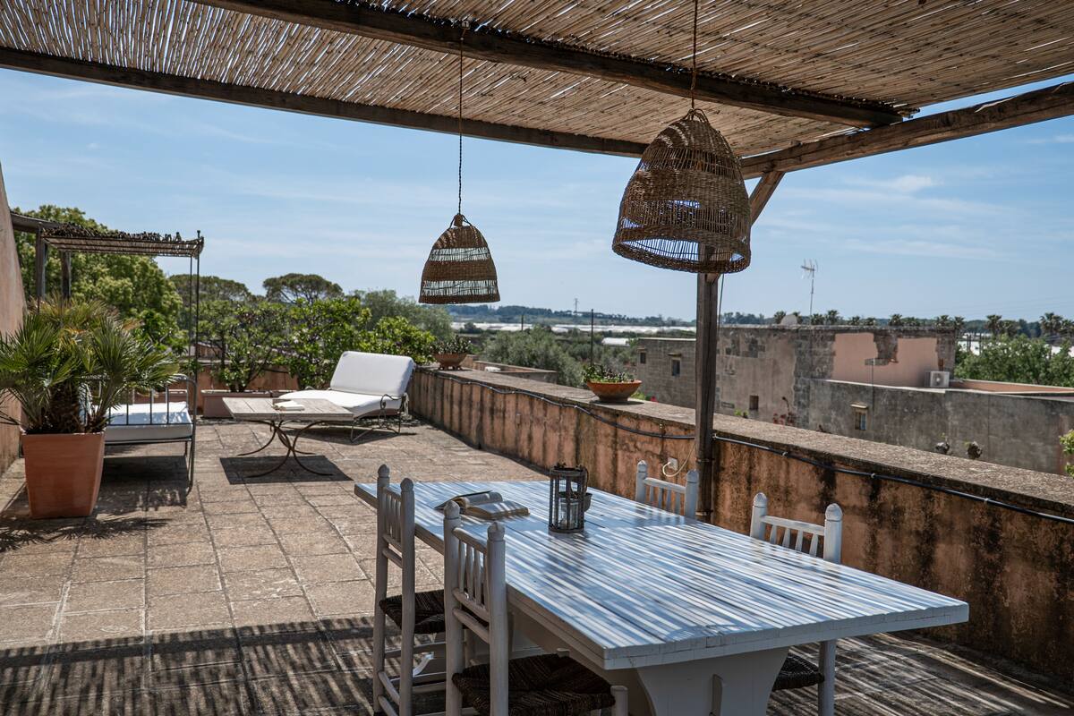A rooftop terrace with a table and chairs, potted plants, hanging wicker lamps, and a view of the cityscape.