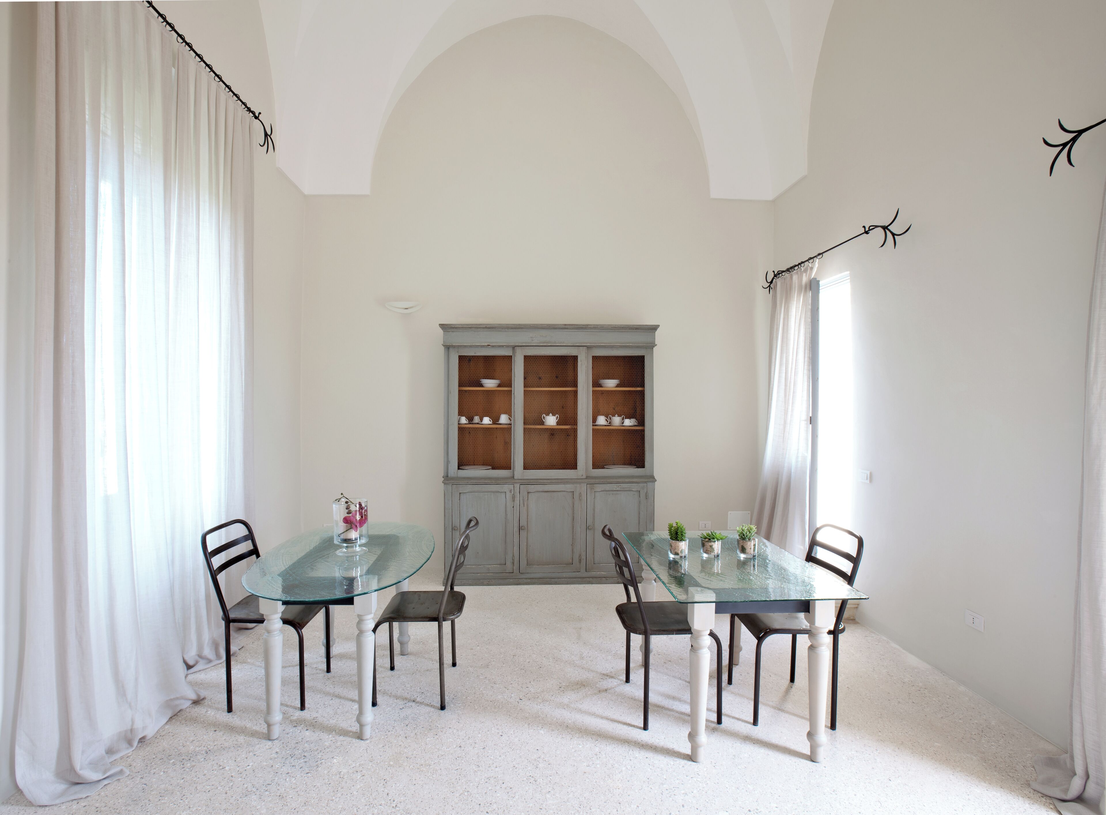 A dining area with a glass table and chairs, a cabinet with dishes, and white curtains.