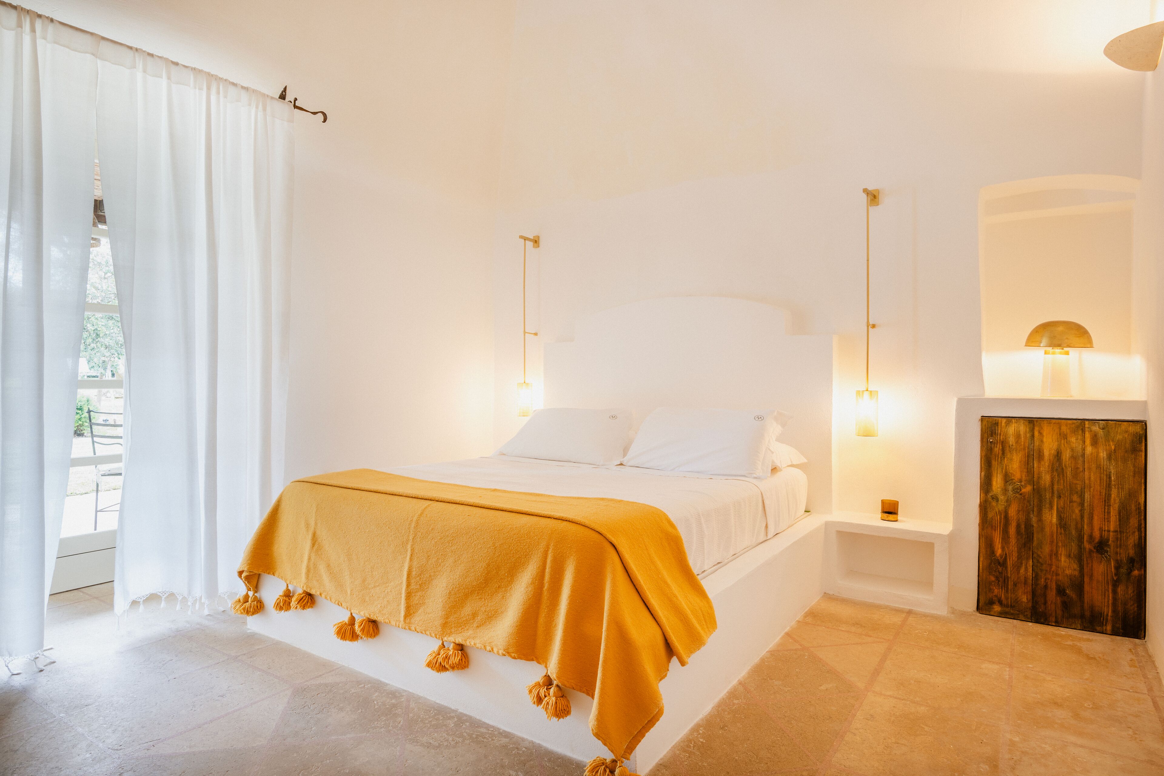 Ein Schlafzimmer mit Bett, weißen Vorhängen, einer Holztür und einem Nachttisch mit Lampe.
