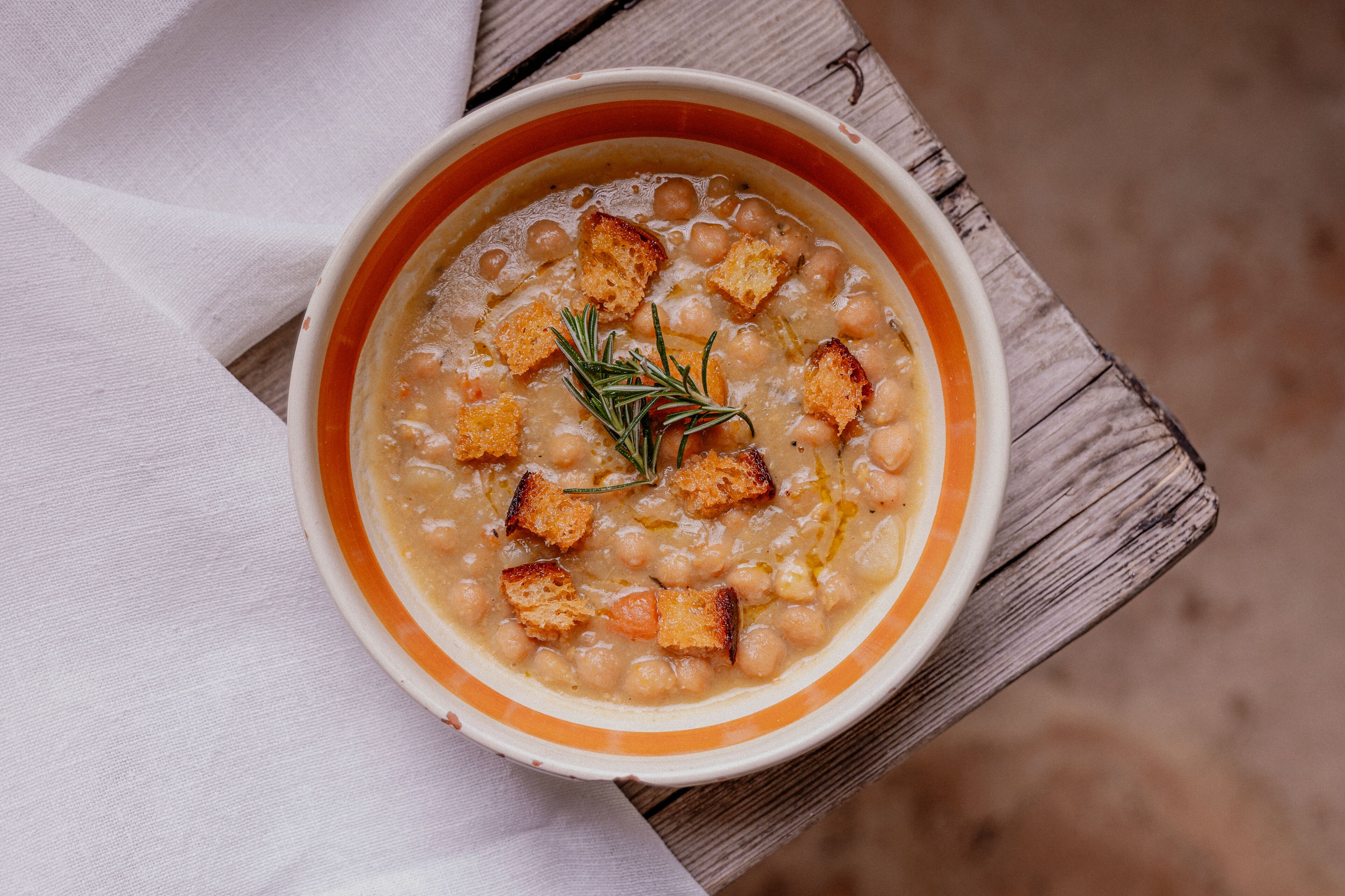 A bowl of soup with croutons and a sprig of rosemary on a wooden surface.