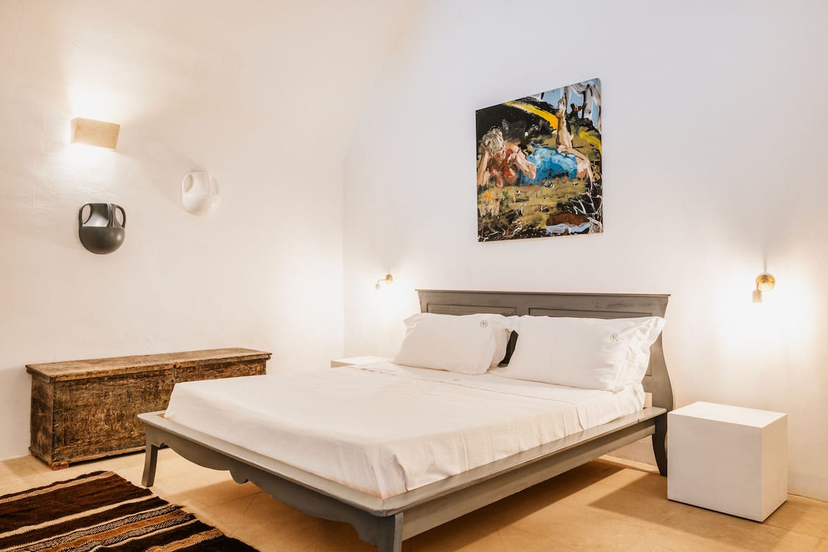 A bedroom with a bed, bedside tables, a nightstand, a painting, and a wall-mounted light fixture.